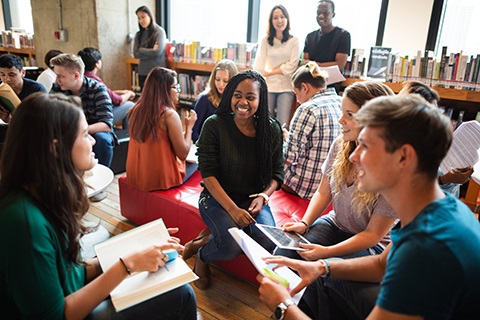 A diverse group of students studying and discussing together in a bright library space filled with bookshelves.