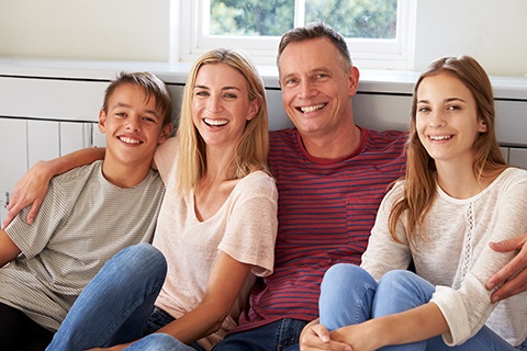 Family of four sitting closely together on the floor in a bright room with a window behind them.