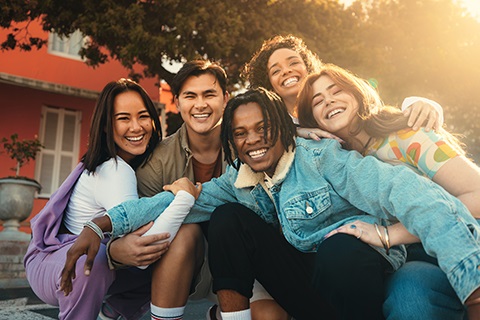 Group of five friends sitting closely outdoors, sharing a warm embrace in front of a red building with sunlight filtering through trees.