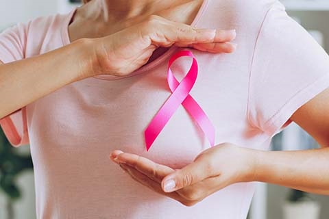 A woman in a pink T-shirt holds her hands up on the top and bottom of a pink ribbon pinned to her chest to highlight breast cancer awareness.