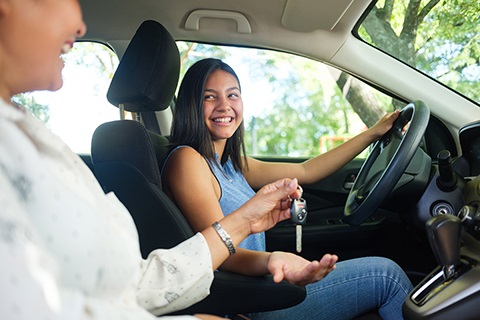 A teenager sits in the driver's seat of a vehicle with her mother in the passenger seat. The mother is placing vehicle keys in her daughter's hand.