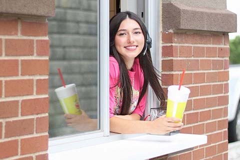 A teenager leans out a drive thru window holding a drink in one hand. She’s wearing a headset and smiling.