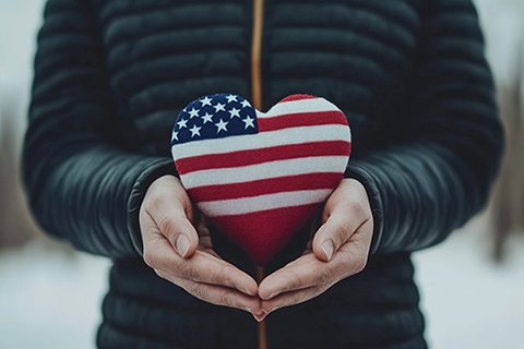 A person wearing a winter coat holds a small heart statue in their hands. The heart is painted to resemble the American flag.