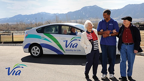 A transit driver assists two elderly passengers walking beside a Via Mobility Transit Service vehicle.