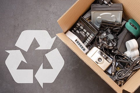 Cardboard box filled with old electronic devices next to a large white recycling symbol on a concrete floor.