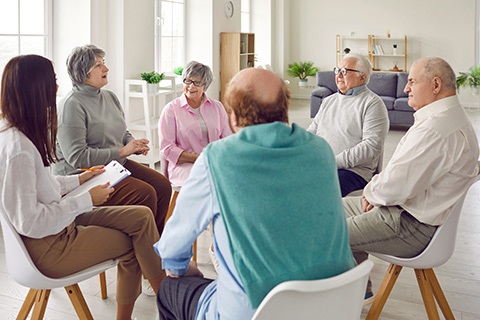 A group of older adult individuals engaged in a supportive discussion in a bright community room setting.