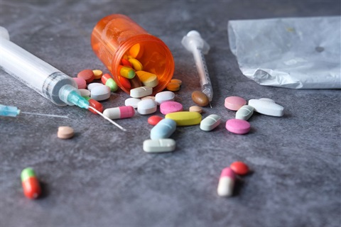 Various colorful pills spilled from an orange prescription bottle next to a syringe and a small plastic bag on a gray surface.