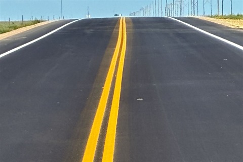 Freshly paved rural road with bright double yellow center lines stretching into the horizon under a clear blue sky.